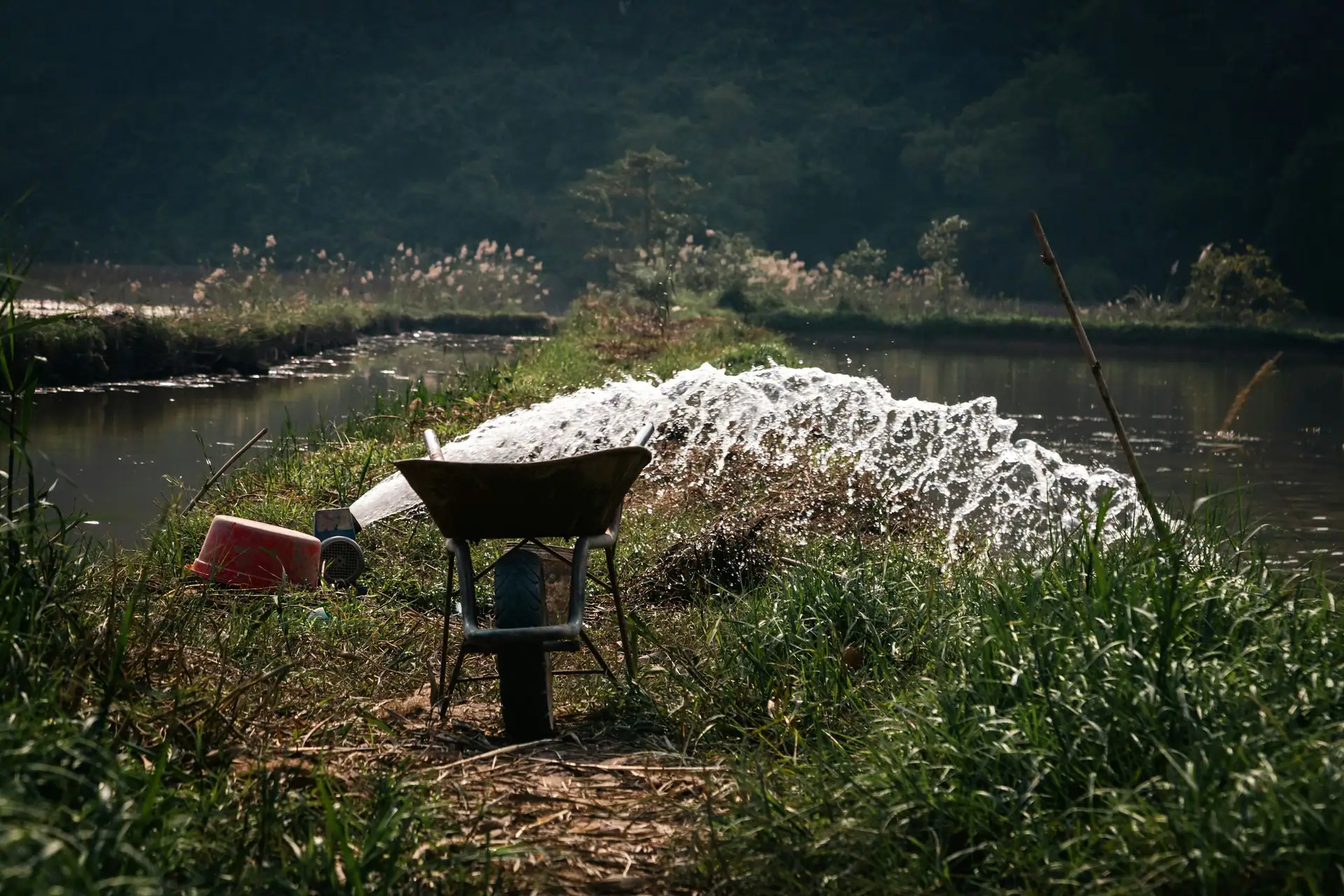 Water jetting from a pipe into a pond, a wheelbarrow on the bank